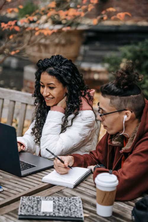 Two people reading documents beside a laptop at an outdoor table.